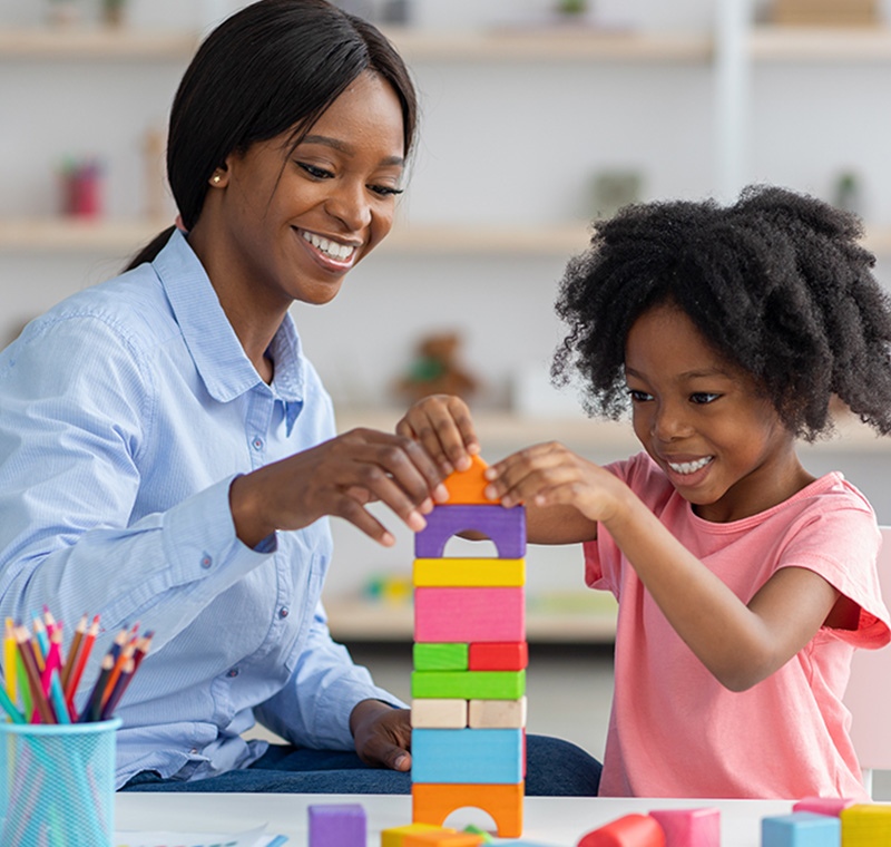 A behavior technician works closely with a child in a therapy-office setting, guiding an activity while reviewing progress charts on a computer screen