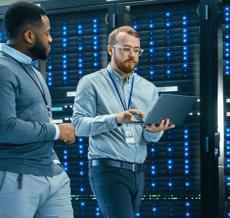 Two IT professionals stand in a data center lined with glowing server racks. One holds a laptop and reviews system performance while the other observes, discussing network operations and infrastructure management.